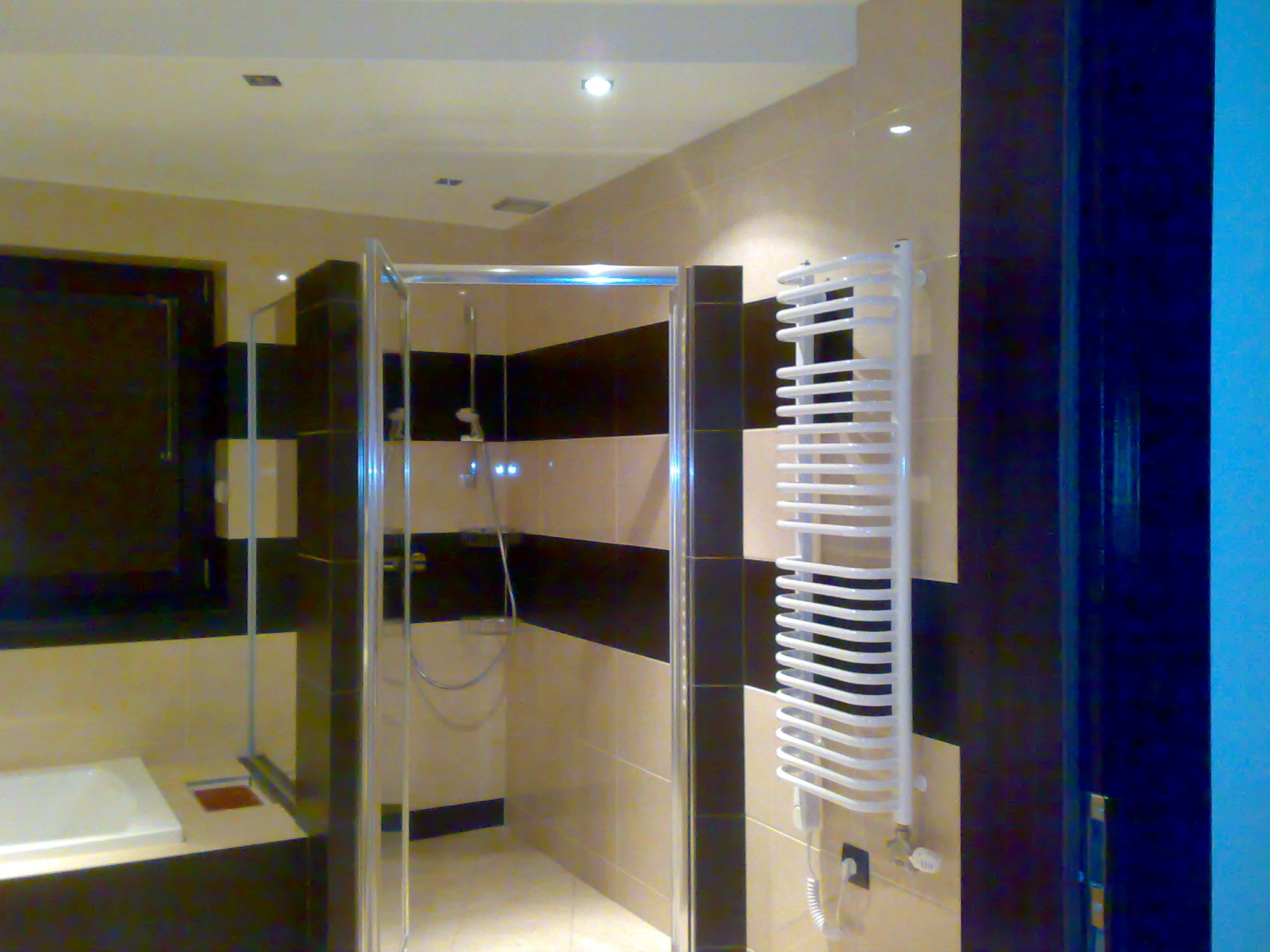 Nowoczesna łazienka z kabiną prysznicową walk-in, wykończona beżowymi i czarnymi płytkami, z białym grzejnikiem drabinkowym i oświetleniem punktowym.